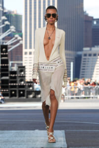 Model walking a city runway in a cream plunging-neckline dress with an embellished belt and decorative cuffs, wearing dark sunglasses.