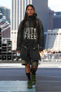 Model wearing an oversized dark denim jacket and shorts on a runway, with a bold 'DUTY FREE' sweatshirt front and green sneakers in an urban backdrop.