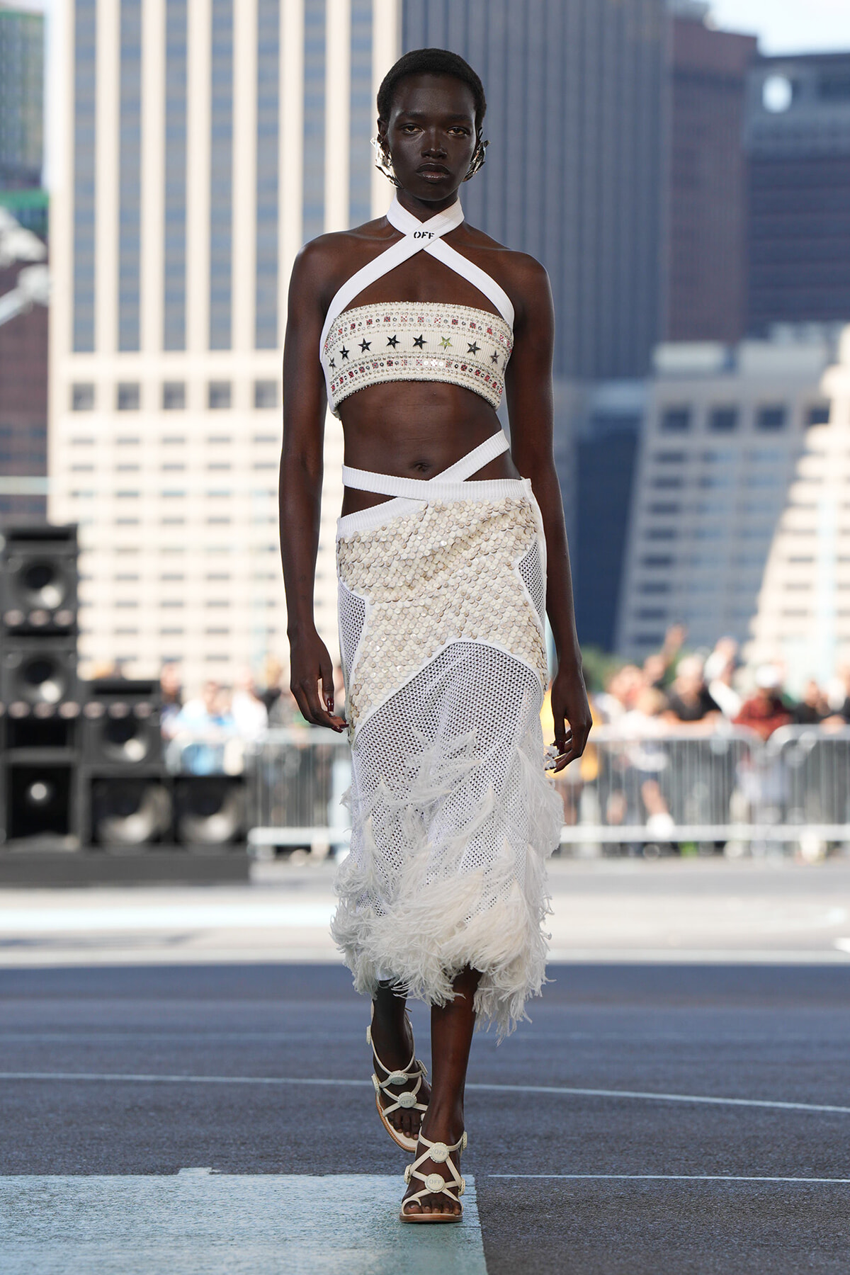 Black model walks a runway in a white embellished halter top and sequined skirt with a feathered hem, urban skyline behind.