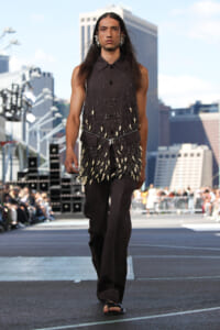 Male model walks a city street runway in a sleeveless dark gray vest densely decorated with shells and beads, paired with black wide-leg pants.