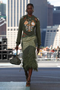 Model wearing an olive-green, cut-out top with skeletal chest graphic and feathered skirt walks a city runway, carrying a small green handbag.