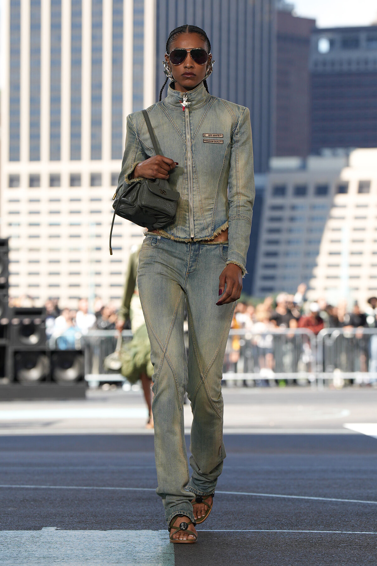 Model wearing a light-wash denim jacket and jeans, tote bag slung over shoulder, sunglasses, walking on an urban runway with skyscrapers in the background.