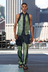 Male model walks a rooftop runway in distressed green denim outfit with layered dark panels and sandals, urban backdrop behind him.
