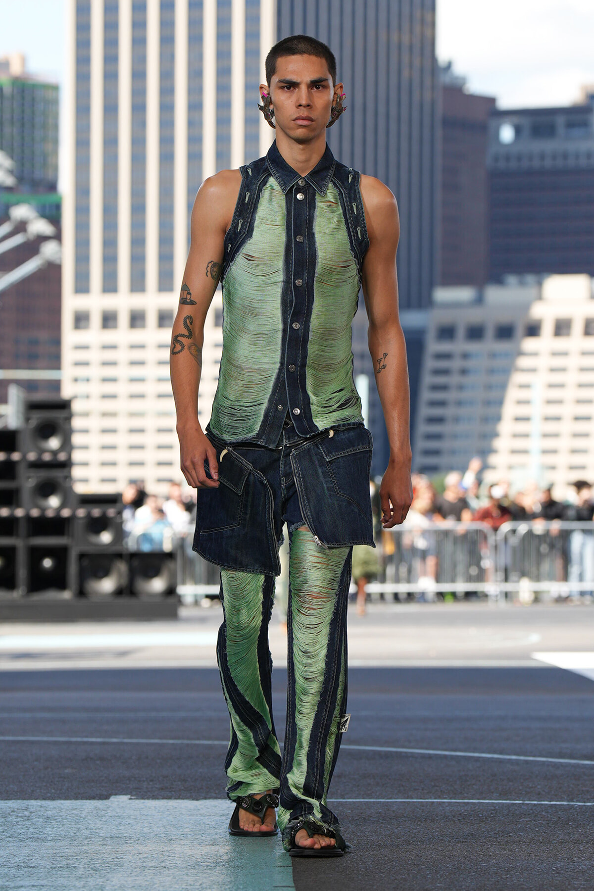 Male model walks a rooftop runway in distressed green denim outfit with layered dark panels and sandals, urban backdrop behind him.