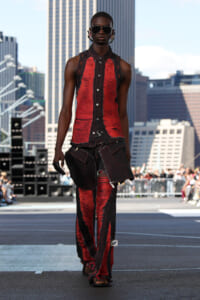 Model walks a rooftop fashion show in a torn red-and-black sleeveless shirt and matching ripped pants, wearing dark sunglasses.