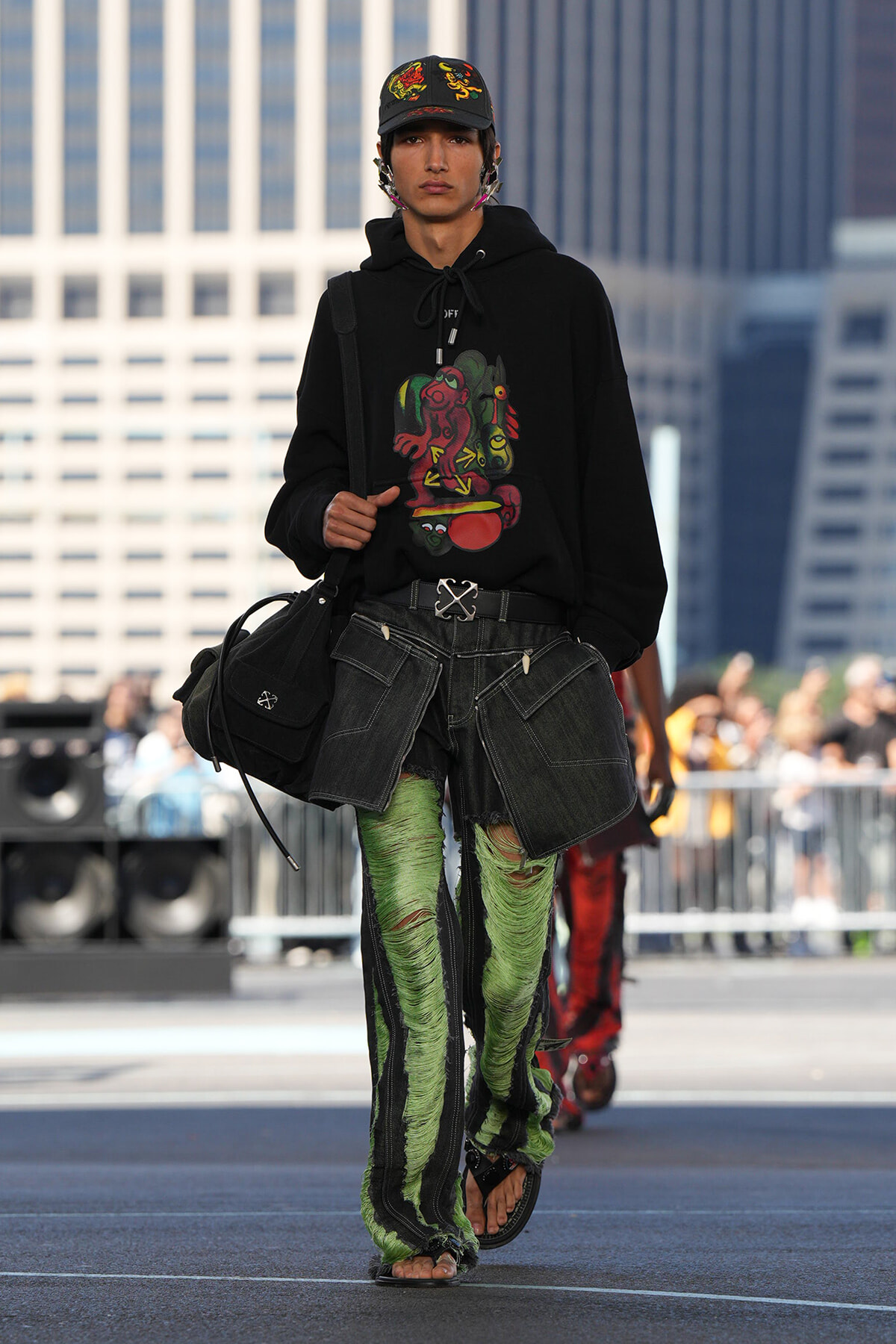 Model walking on an urban runway wearing a black hoodie with a colorful graphic, green distressed jeans, and sandals, carrying a black bag and wearing a patterned cap.