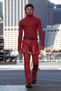 Male model walking a city runway in a red knit hooded top and matching red leather pants with zippers and pockets; urban skyscrapers in background.