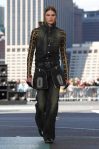 Model walking a city runway in a layered black denim vest over a yellow-plaid hooded top, with oversized cargo-style pants and gloves.