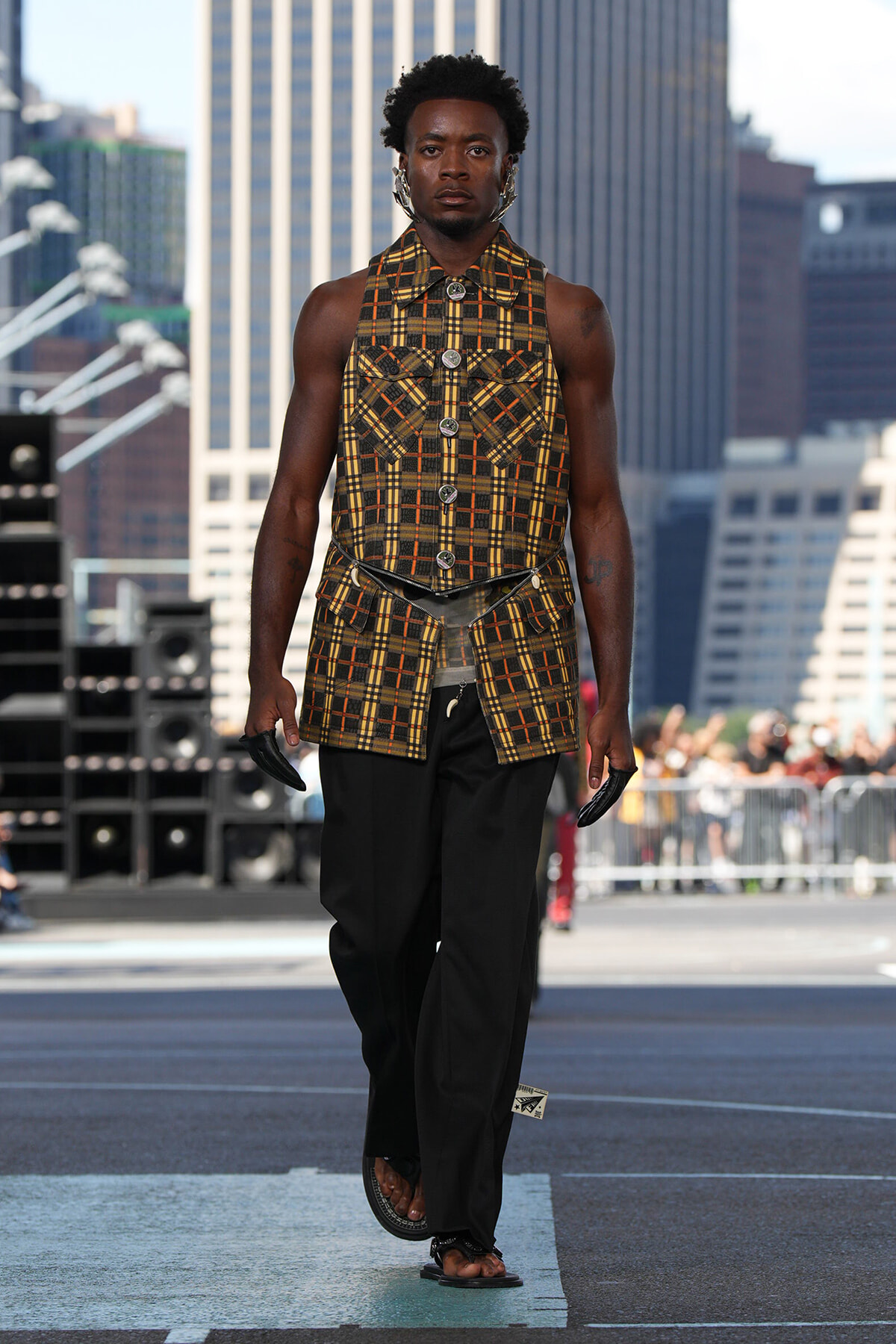 Male model walking on a city street runway in a brown plaid sleeveless jacket with large buttons, paired with black wide-leg pants and sandals.