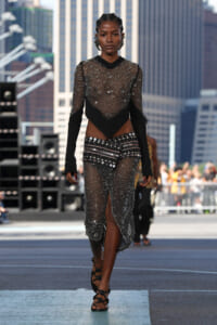 Model walks down urban runway in a black mesh, sequined bodysuit with a high-low skirt and strappy sandals.
