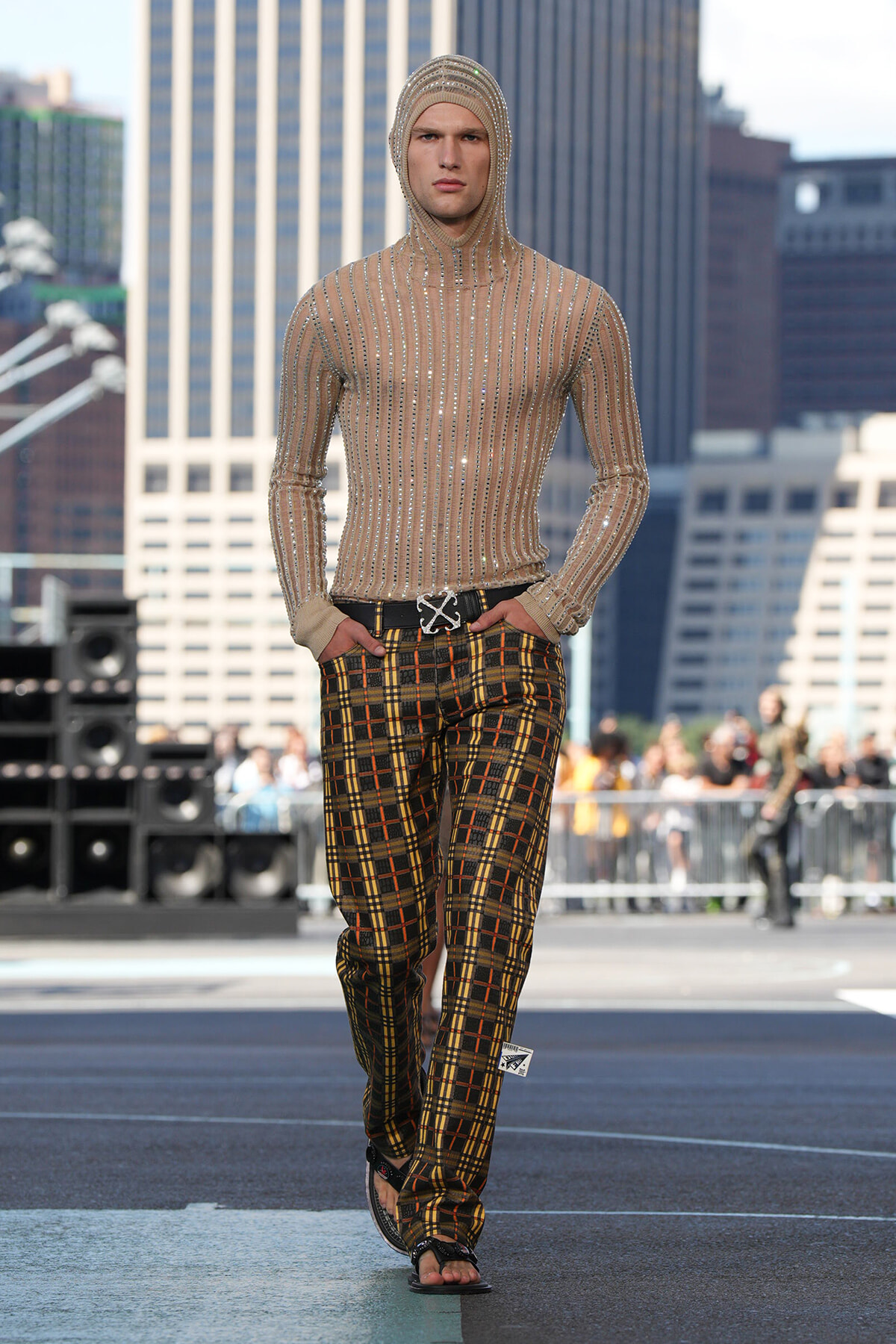 Male model walking a city rooftop runway in a beige sequined hooded top and yellow plaid trousers with a black belt; urban skyline behind him.