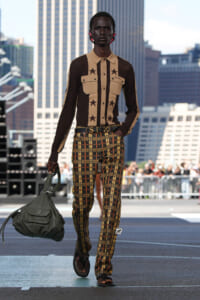 Male model walks a city rooftop runway in a beige and brown star-pattern shirt with plaid yellow trousers, carrying a large gray bag.