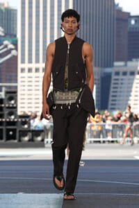 Male model walking on a city runway in a layered black quilted vest over a plaid shirt, black pants, and flip-flop sandals; urban skyline backdrop and spectators in the background.