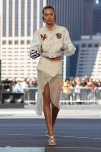 Model on a city runway wearing a cream varsity jacket with patches and a textured wrap skirt.