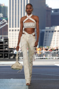 Model walks a city runway in a white crisscross halter top with star-embellished band and light beige cargo pants, carrying a cream handbag.