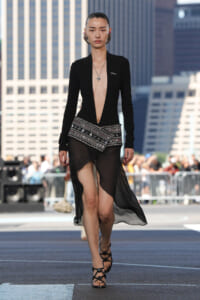 Model walking a runway in a black plunging gown with a jeweled belt and sheer asymmetrical skirt, urban cityscape in the background.