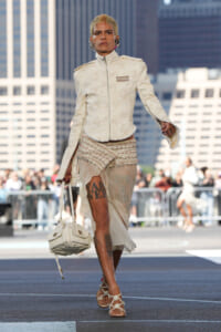 Runway model in a white textured jacket and beaded wrap skirt walks outdoors in a city street, carrying a white handbag and wearing beige heeled sandals.