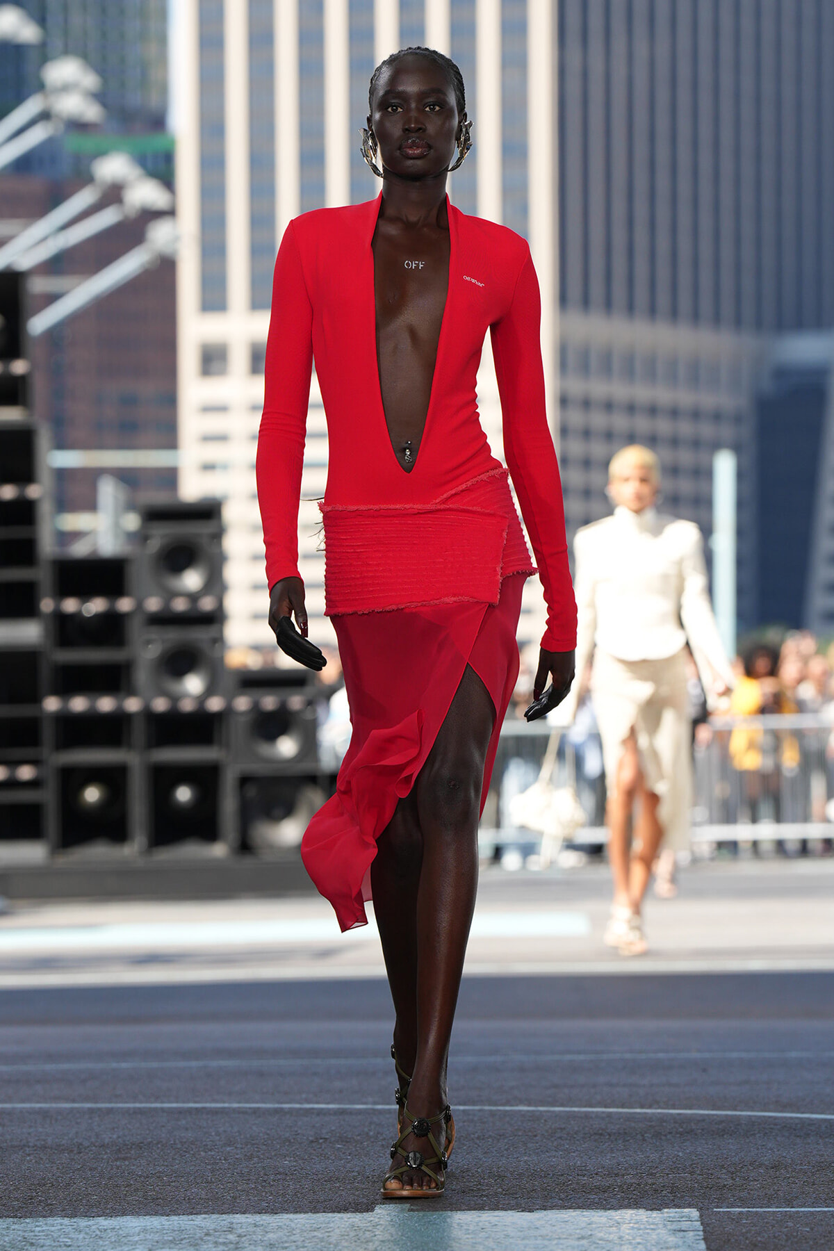 Runway model in a form-fitting red dress with a deep plunging neckline and high slit, walking on an city street-themed catwalk with a blurred urban background.