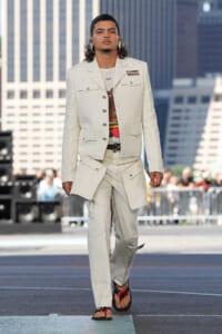 Male model walking a city-front runway in a cream-colored utility suit with oversized pockets, graphic tee, and belt, urban skyscrapers in the background.