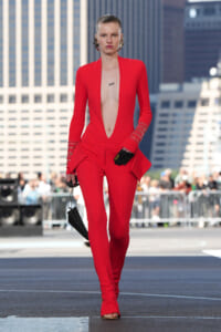Model walking a street runway in a bold red jumpsuit with a plunging neckline and embellished cuffs against a city backdrop
