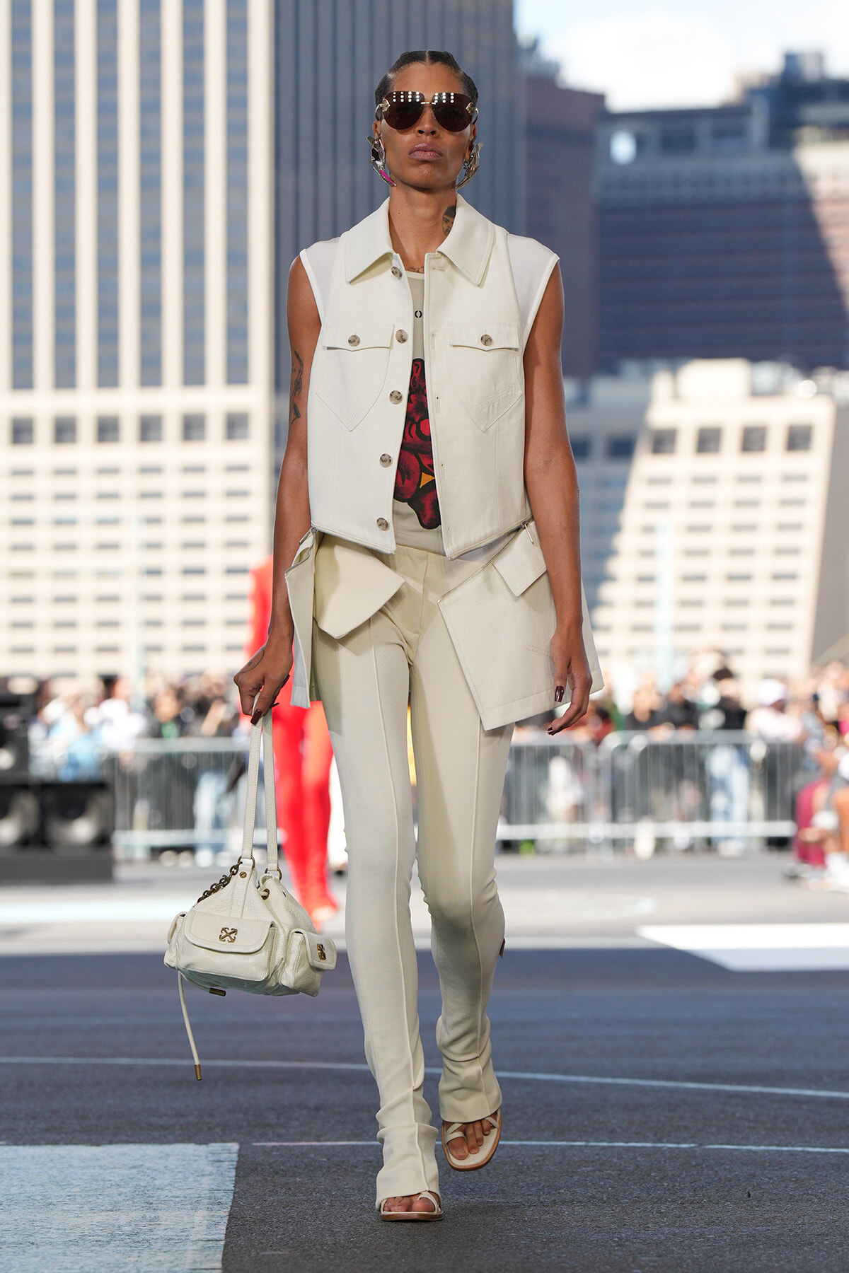 Model walks a runway in a cream sleeveless vest and matching slim cargo pants, carrying a cream handbag against a city skyline backdrop.