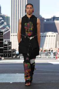 Male model on a city runway wearing a black layered sleeveless hoodie with zippers and colorful monster prints on top and pants, urban skyline backdrop