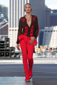 Model walking a city runway in red pants and a sheer black top with red star panels, against skyscrapers in the background.