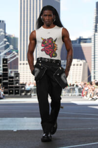 Runway model walking in a white sleeveless shirt with a red cartoon demon graphic and black cargo pants on a city street backdrop.