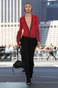Model walks a city runway wearing a red zip-front mesh bodysuit, black tailored pants, and a small black handbag.
