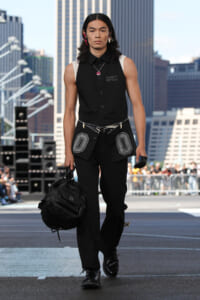 Male model walks an urban runway in an all-black outfit: sleeveless shirt, pants, belt with decorative pockets, pink earrings, and a large black duffel bag in hand.