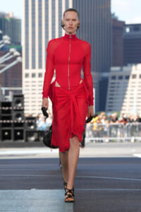 Model walking a rooftop runway in a red zip-front top and draped red skirt with a waist knot; black gloves and strappy heels, city buildings behind.