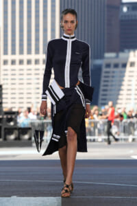 Model walks a runway in a navy black and white zip-front jacket with matching layered skirt and geometric handbag, city skyline behind.