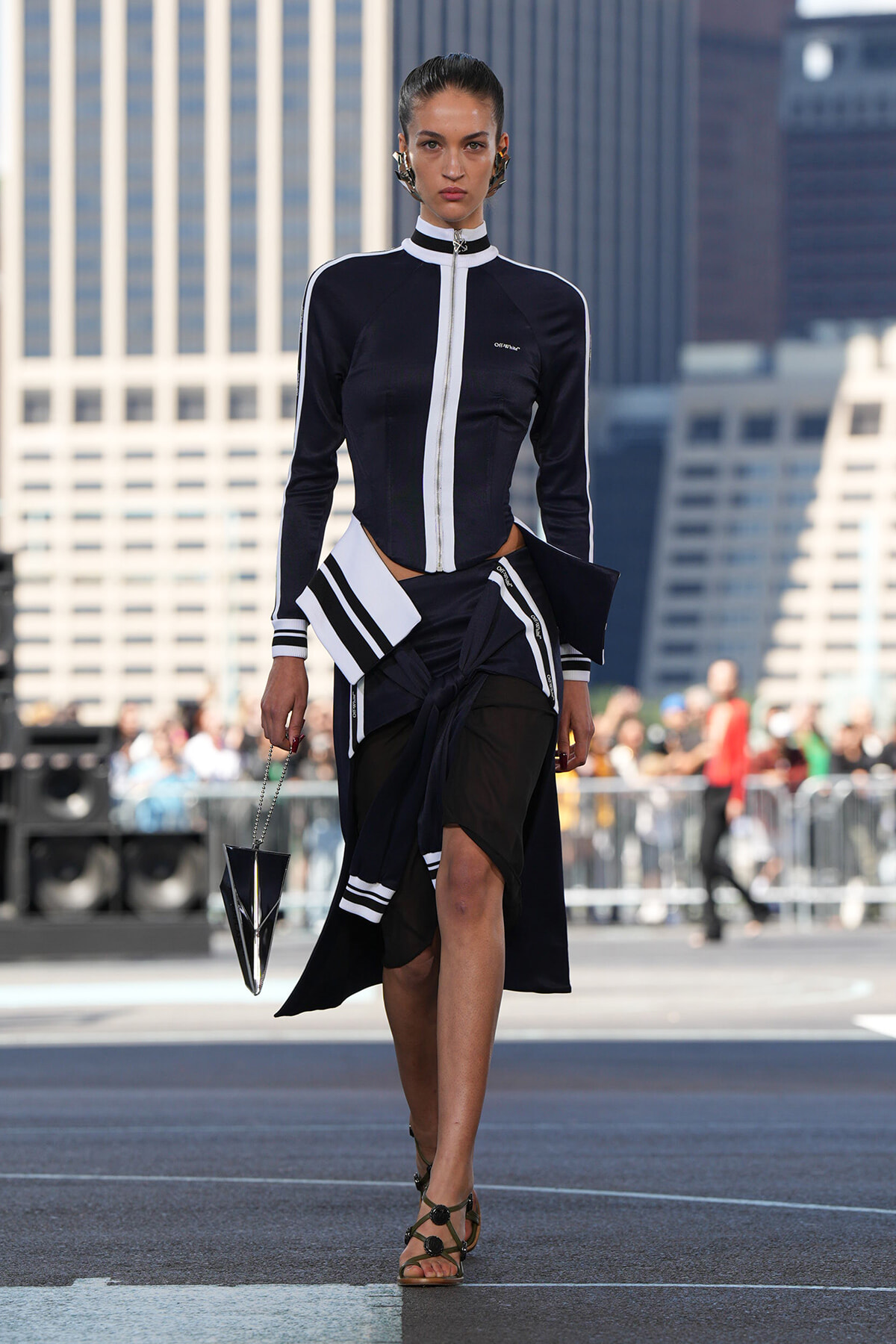 Model walks a runway in a navy black and white zip-front jacket with matching layered skirt and geometric handbag, city skyline behind.