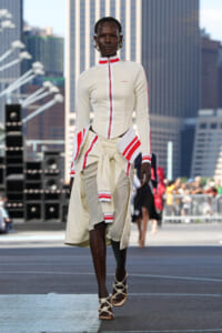 Model walks a street-fashion runway in a cream outfit with red stripes and a high collar, showcasing a structured zip-front top and layered skirt with a tied sweater.
