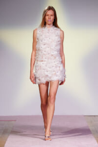 Model walking a runway in a sleeveless white floral-textured mini dress with pinkish sandals, on a soft pink runway backdrop.