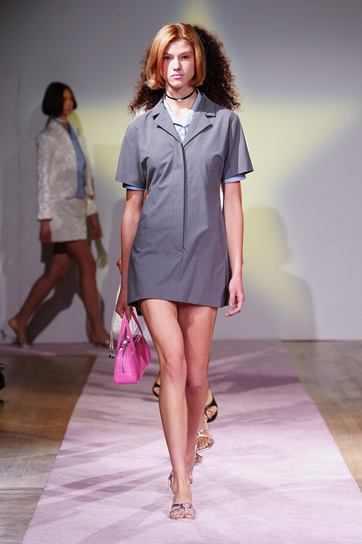 Model walks the runway in a gray short-sleeve dress, pink handbag, and strappy sandals under pastel lighting.