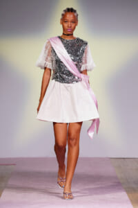 Model on a runway wearing a silver sequin top with sheer lace sleeves, a pale pink sash, and a short pink skirt.