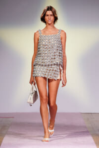Model walks the runway in a silver floral-beaded sleeveless mini dress, white sandals, and a white handbag under bright stage lights on a pink runway.