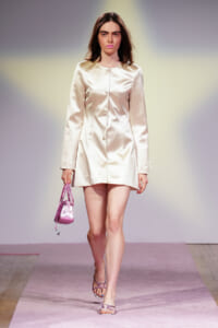 Model on a fashion show runway wearing a cream satin coat-dress with a pink handbag and strappy sandals on a pink catwalk surface.