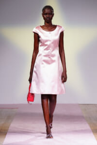 Model in pale pink satin A-line dress walking the runway, holding a small pink bag and wearing silver sandals.