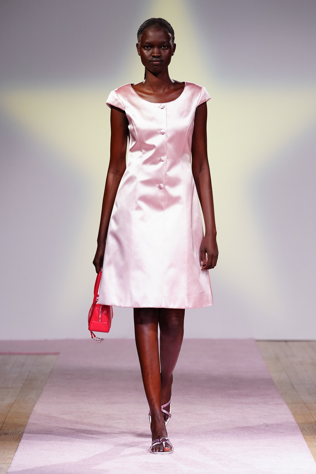 Model in pale pink satin A-line dress walking the runway, holding a small pink bag and wearing silver sandals.