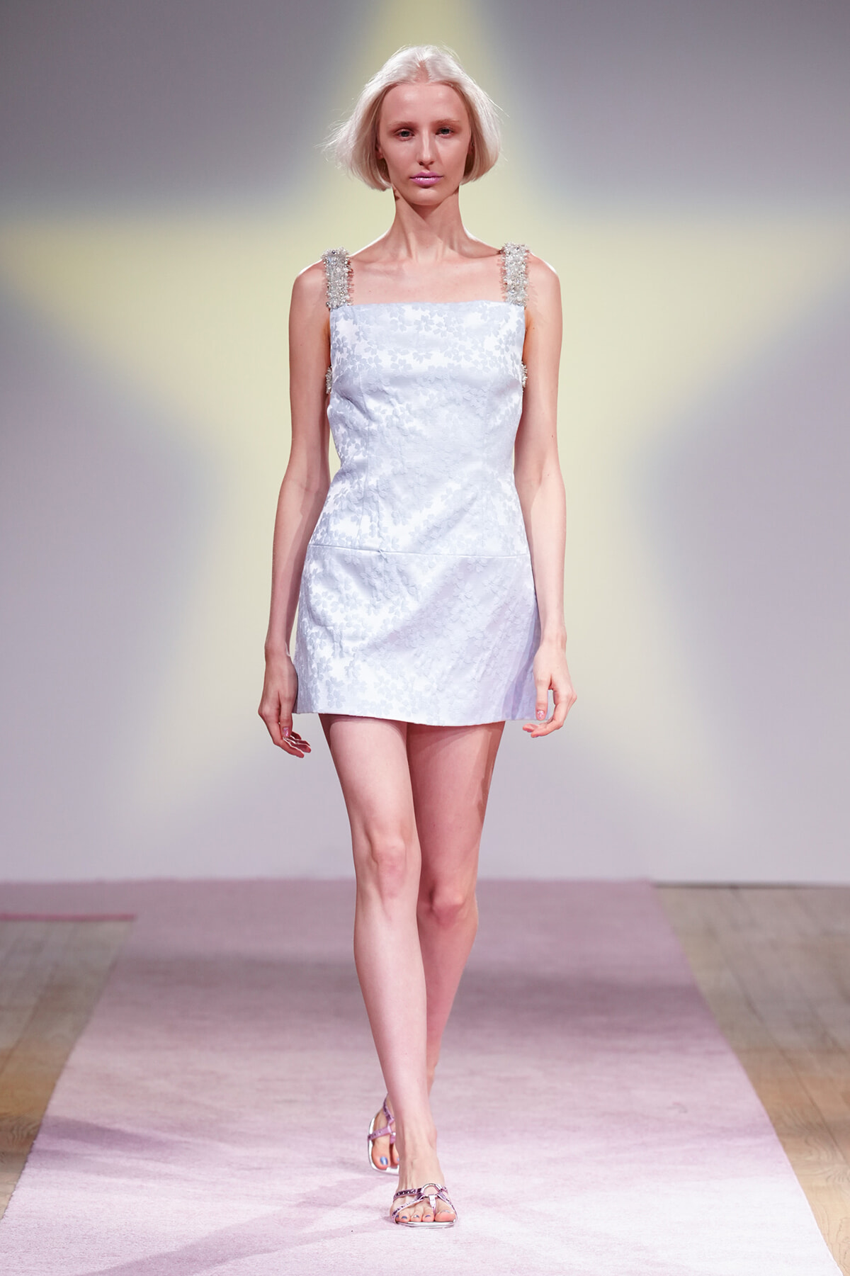 Model walks the runway in a pale blue floral mini dress with beaded straps and silver sandals.