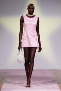 Female model walking a pink runway in a pale pink lace mini dress with a diagonal sash, paired with a pearl choker and a white floral clutch held in her left hand.