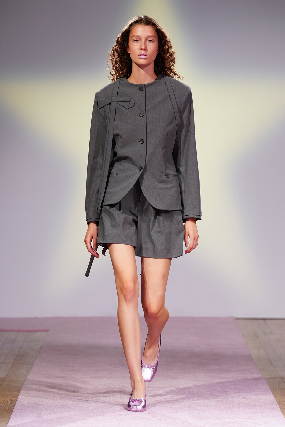 Model walking a fashion runway in a tailored gray suit with a short matching bottom and lavender flats.