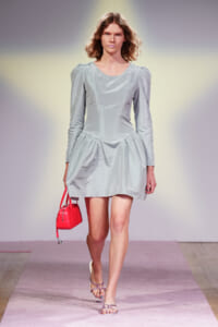 Model walking a runway in a pale blue, structured dress with a full skirt, carrying a bright red handbag on a pink catwalk.