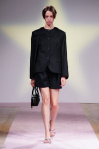 Model on a runway wearing a black tailored blazer and matching shorts, holding a small black handbag.