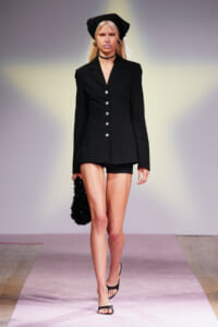 Model walks runway in a tailored black blazer with front buttons, black shorts, headscarf, and choker, carrying a black textured bag.