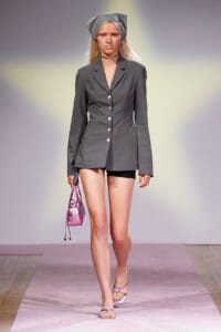Model walks the runway in a gray blazer-style dress with a buttoned front, pairing black shorts, a gray hat, a pink handbag, and lilac strappy sandals.