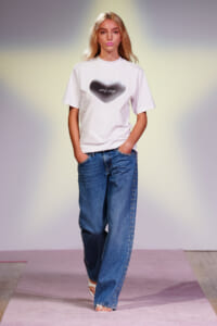 Model walks the runway in a white T-shirt with a black heart graphic and loose blue jeans studded along the sides.
