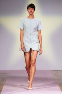 Runway model wearing a light blue zip-front short-sleeve dress walking on a pink catwalk.